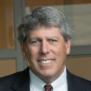 A middle-aged man with gray hair, wearing a dark suit, white dress shirt, and maroon patterned tie, smiles at the camera. He is standing indoors in front of a window with blurred background.