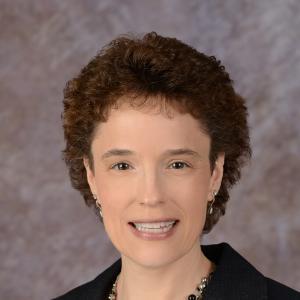 A person with short, curly brown hair and wearing a dark blazer smiles against a blurred grayish background. They have pearl earrings and a necklace with a small pendant.