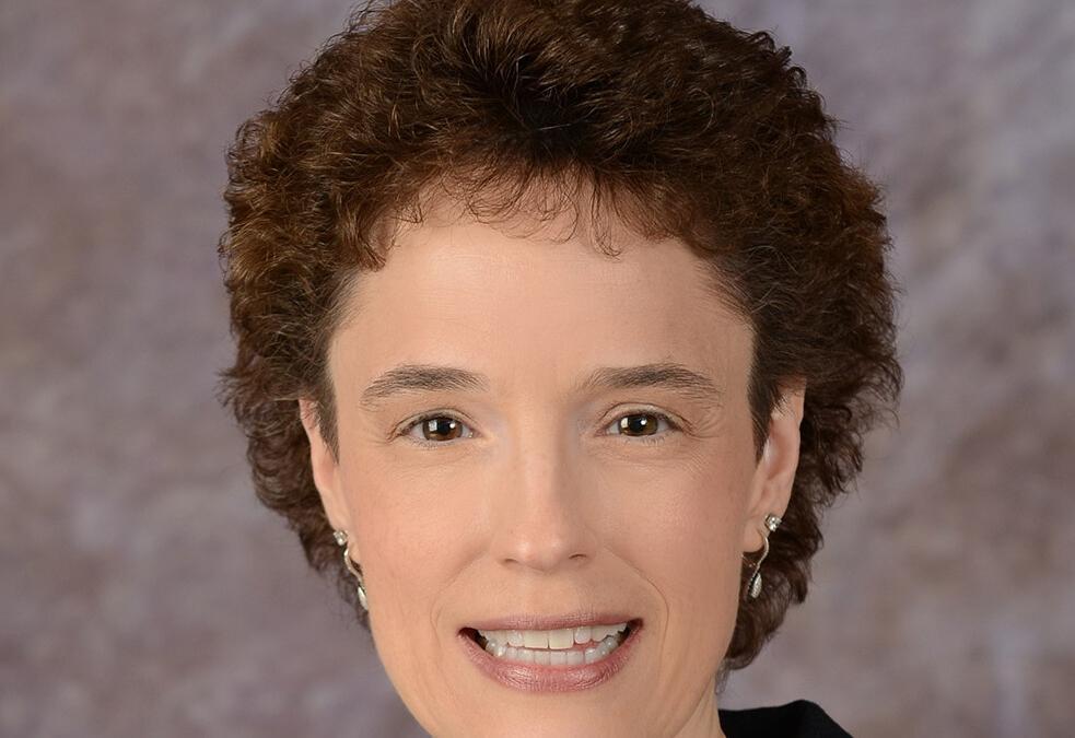 A person with short, curly brown hair and wearing a dark blazer smiles against a blurred grayish background. They have pearl earrings and a necklace with a small pendant.