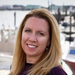 A woman with long blonde hair smiles at the camera, standing outside near a waterfront with boats and docks in the background. She wears a dark top, and the sky is overcast.