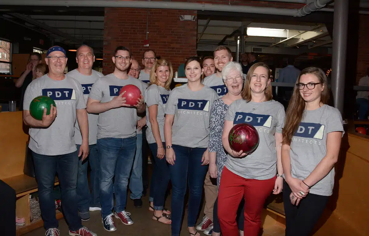 A group of people wearing matching gray t-shirts with the PDT logo stand together in a bowling alley. Several are holding colorful bowling balls and smiling at the camera. The setting has a casual and fun atmosphere with brick walls in the background.