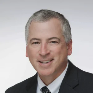 A smiling man with short gray hair wearing a dark suit, white shirt, and polka dot tie is posed against a plain white background.