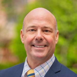 A bald man is shown outdoors, smiling at the camera. He is wearing a dark blue suit jacket, a white and blue checkered shirt, and a yellow and blue striped tie. The background is blurred, with green foliage and some indistinguishable structures.