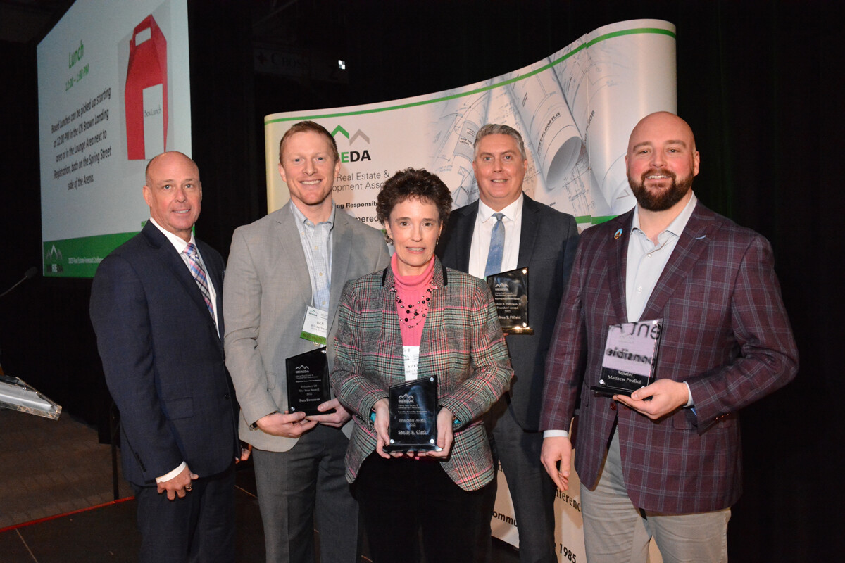 Five people are standing in a row indoors, holding awards and smiling at the camera. They are dressed in business attire, with a backdrop featuring a display about real estate and economic development.