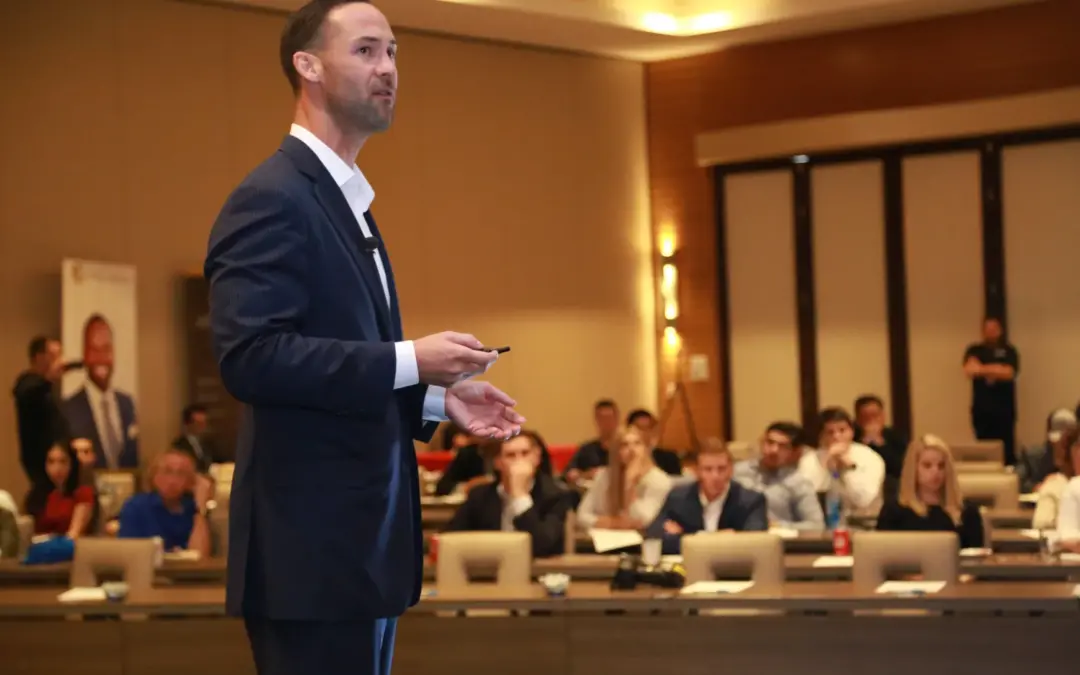 A man in a suit stands on stage holding a pointer, giving a presentation to a seated audience in a conference room. The audience is composed of several people attentively listening and taking notes on their devices. Large windows and a banner are seen in the background.