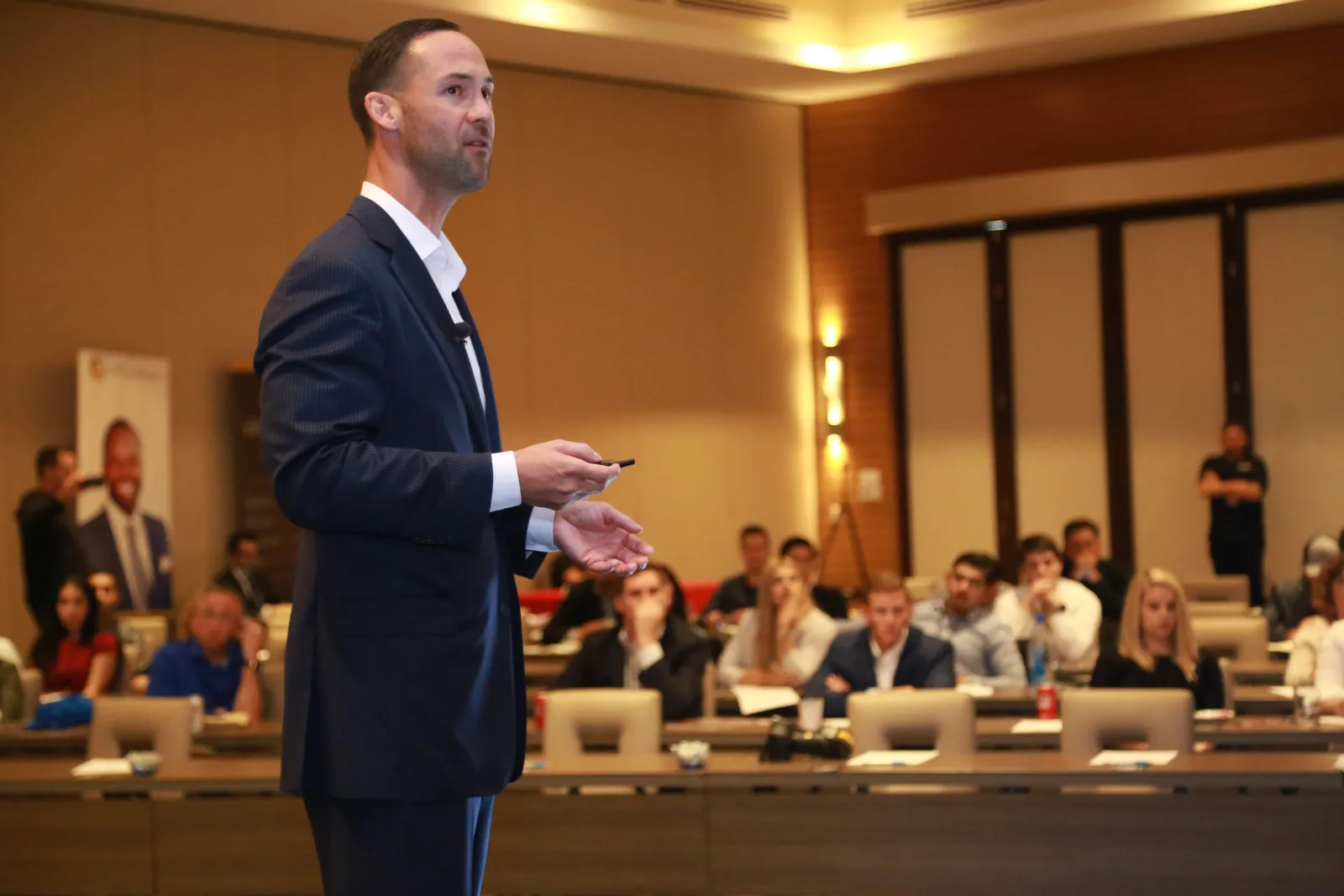 A man in a suit stands on stage holding a pointer, giving a presentation to a seated audience in a conference room. The audience is composed of several people attentively listening and taking notes on their devices. Large windows and a banner are seen in the background.