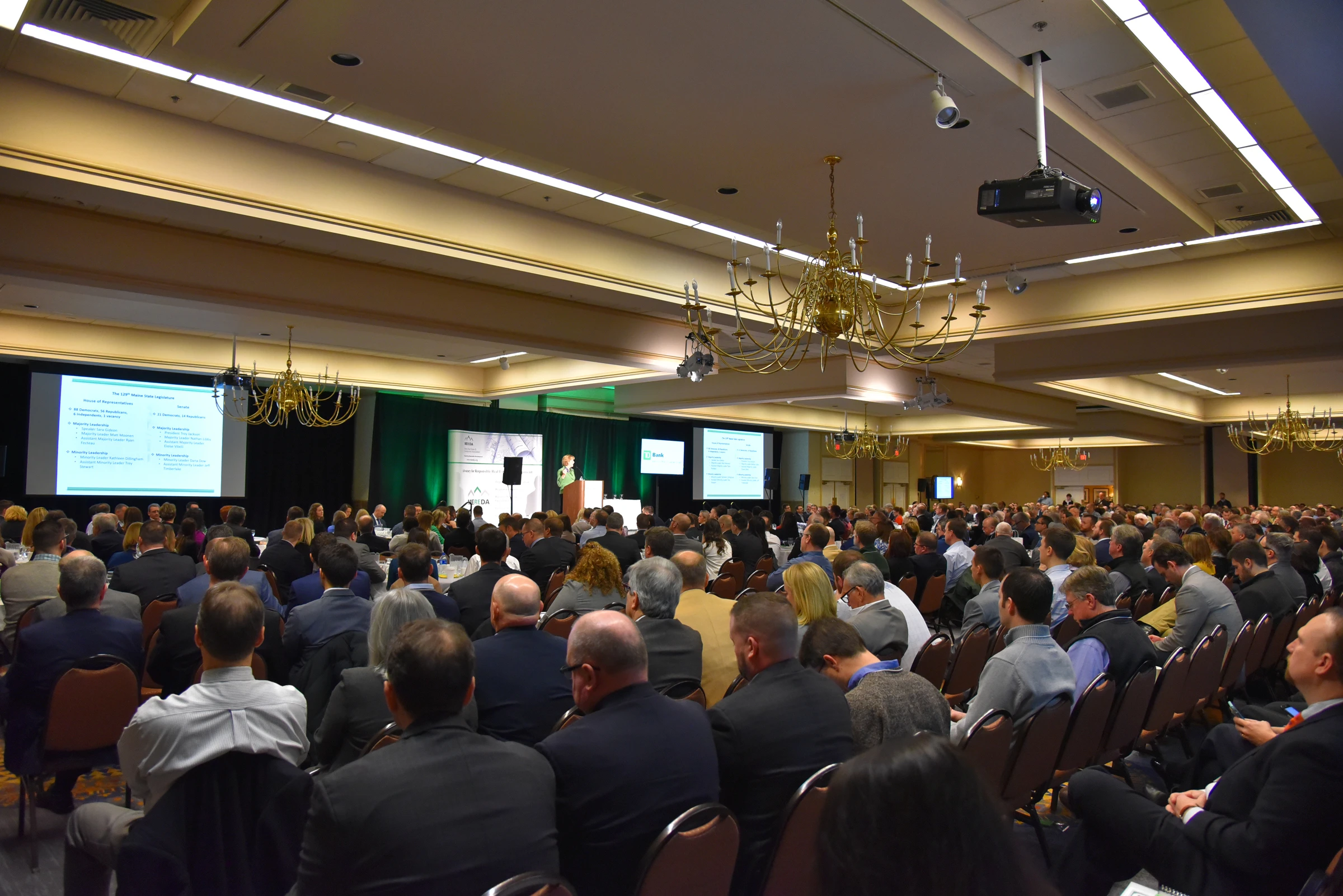 A large conference room filled with seated attendees facing a stage. The stage has a person presenting a slideshow displayed on multiple screens. The room's chandeliers and ceiling lights are visible, and the audience is attentive.