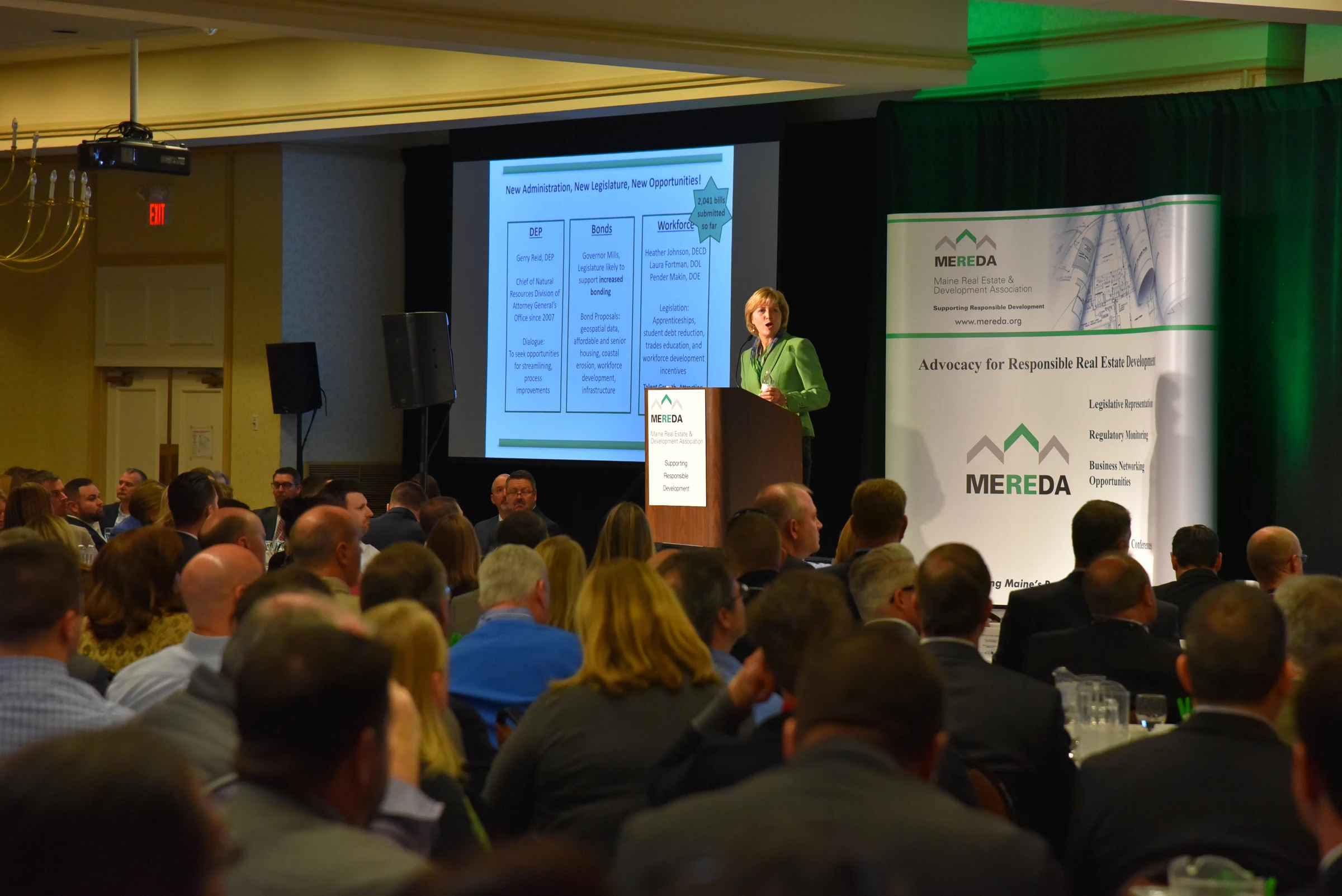 A woman addresses a large audience in a conference room. She stands at a podium with the MEREDA logo, with a presentation slide projected behind her.