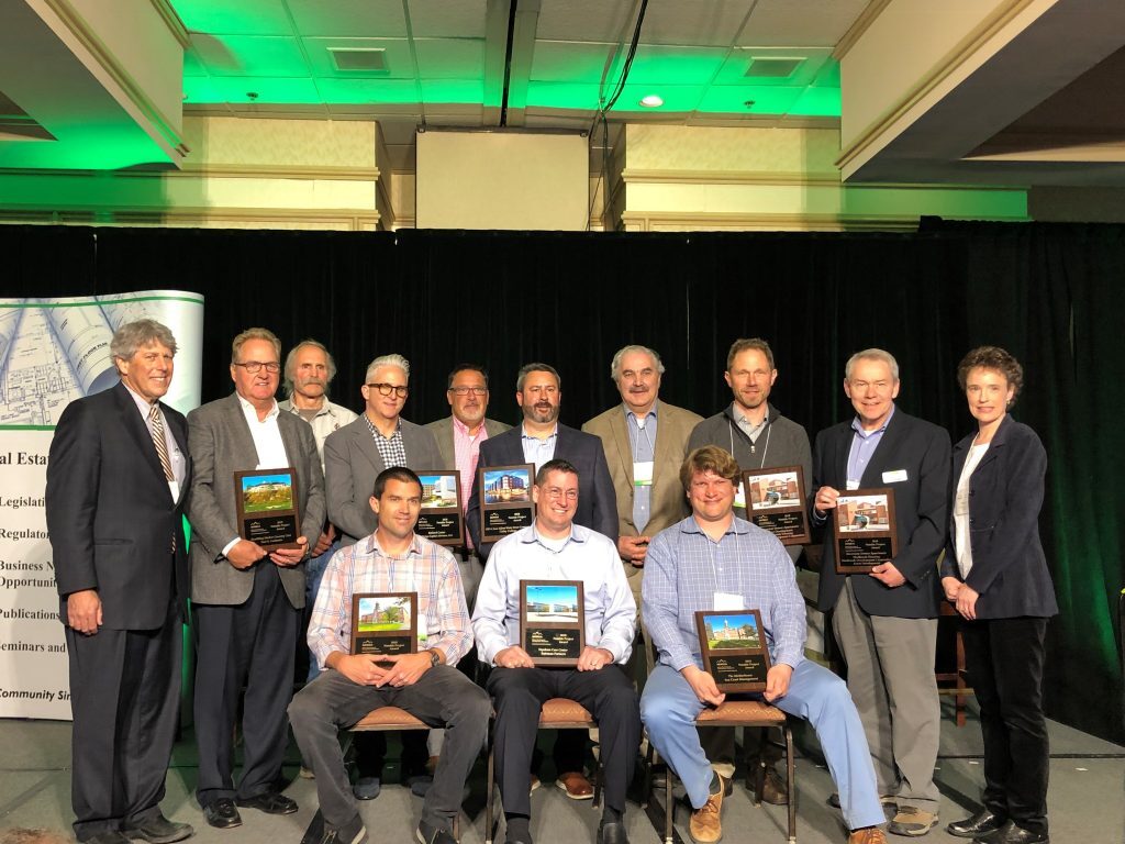 A group of 14 people posing for a photo on a stage. Eleven are standing at the back, and three are seated in front. Several people are holding plaques. This is from the Maine Real Estate & Development Association Awards Top 6 Notable Projects of 2018.