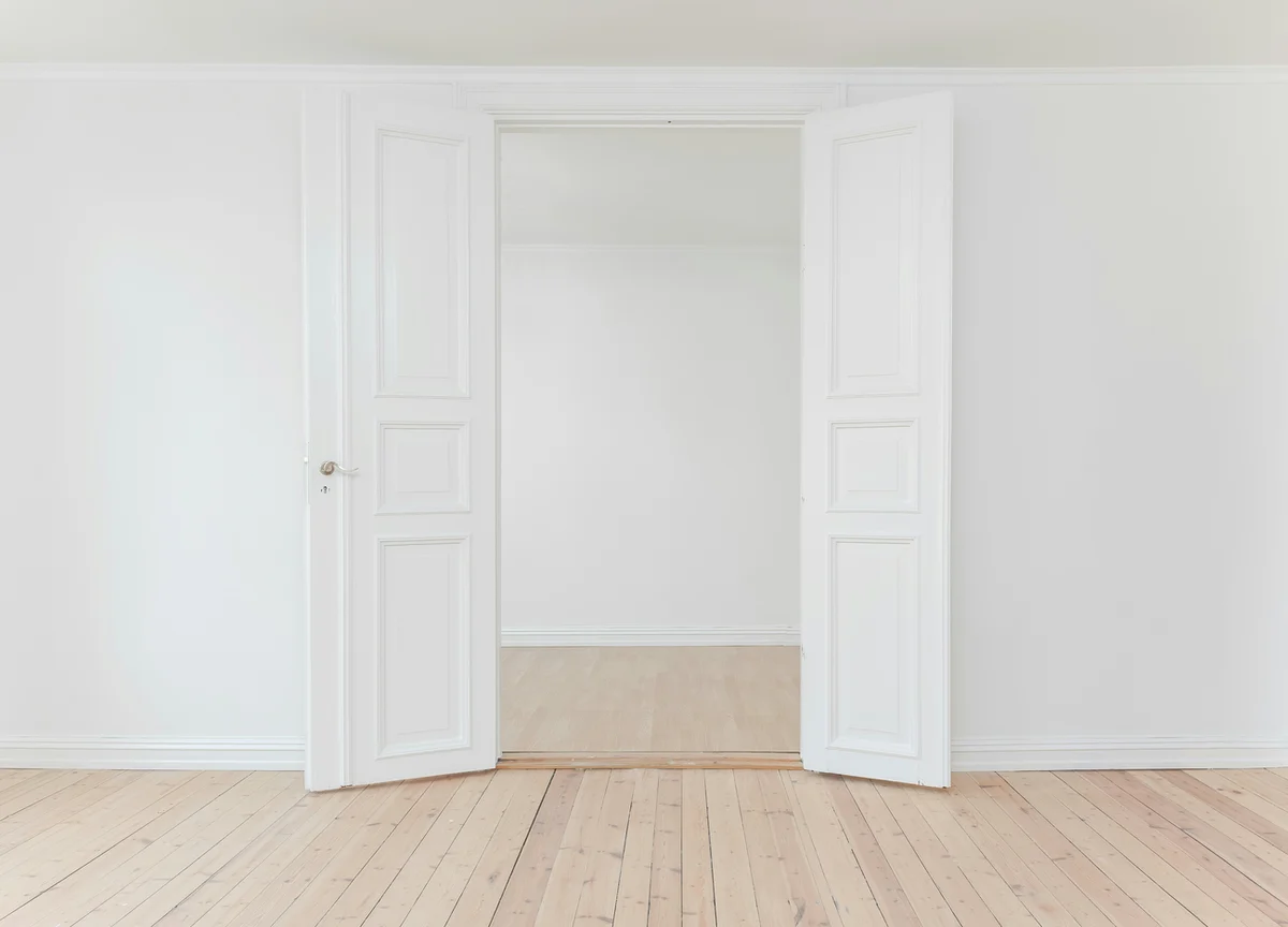 A minimalistic, empty room with light wooden flooring and white walls. The room features double-door white panels that are partially open, leading to another empty space with identical decor. The overall aesthetic is clean and modern.